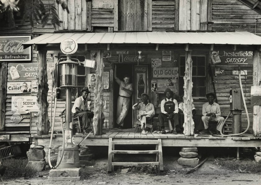 Dorothea Lange (American, 1895-1965) 'Country store on dirt road. Sunday afternoon. Note the kerosene pump on the right and the gasoline pump on the left. Rough, unfinished timber posts have been used as supports for porch roof. Black men are sitting on the porch. Brother of store owner stands in doorway, Gordonton, North Carolina' July 1939, printed later